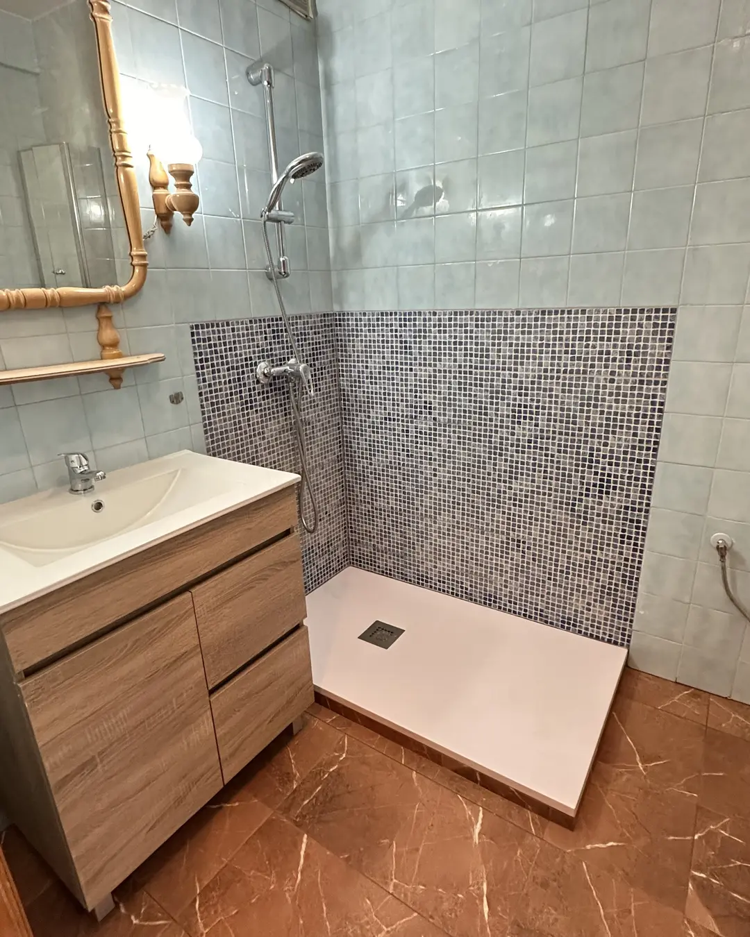 El mismo rincón terminado: plato de ducha extraplano, frente de mosaico gris y mueble de lavabo nuevo, conservando el azulejo original