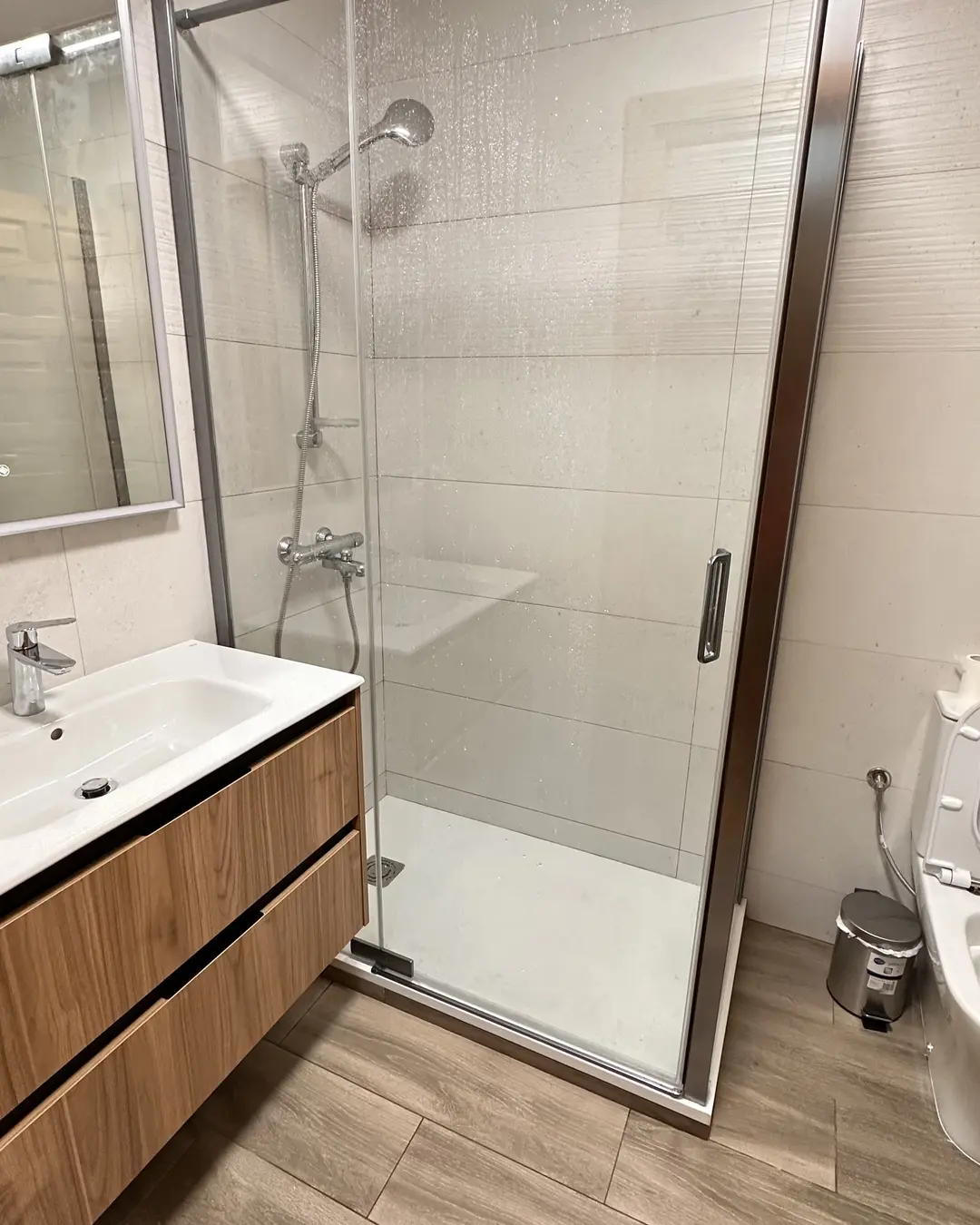 El mismo baño reformado: ducha con mampara de cristal, mueble suspendido de madera y suelo porcelánico efecto roble